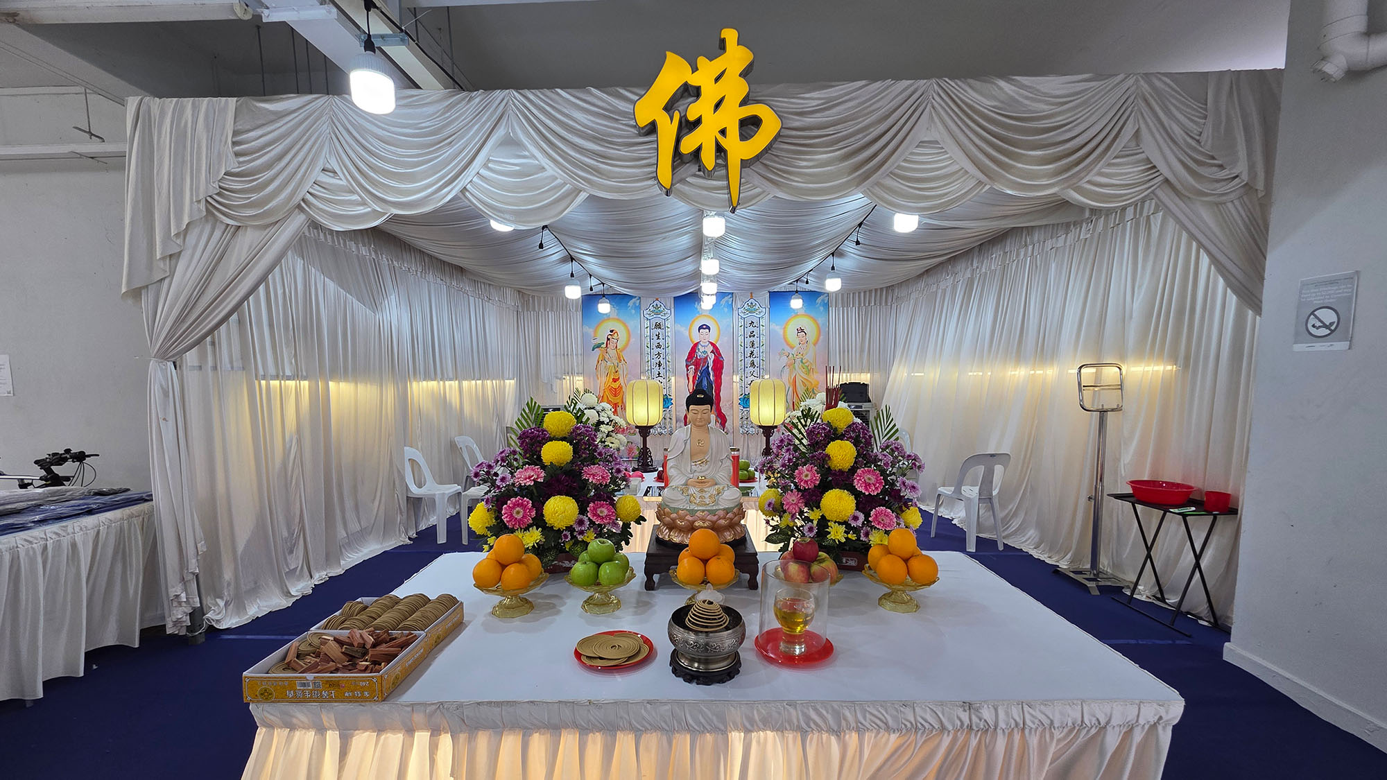 A Buddhist funeral altar setup in Singapore with the Buddha character, fruit and incense offerings, and chrysanthemum arrangements